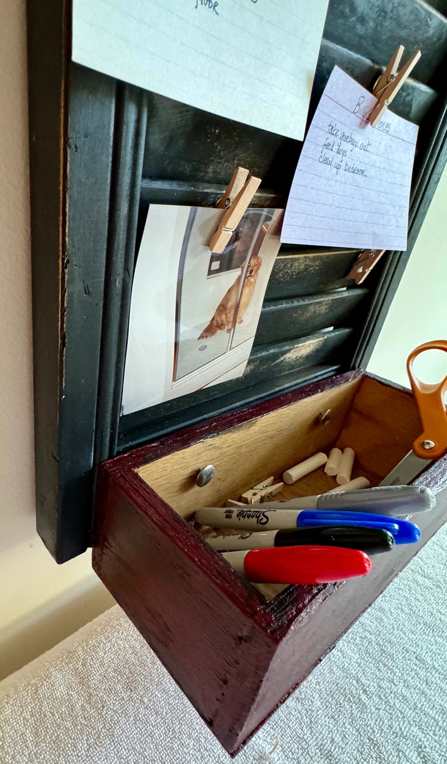 close up of bottom of shutter showing wooden container displayed with chalk, pens and scissors.  Can see clothes pins with photos and cards