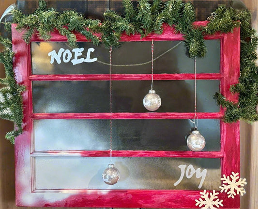 4 retangular horizontally paned old window painted rustic red with faux snow on window panes and words Noel and Joy painted silver.  garland on top and half-way down sides of window and 3 silver balls hanging from top